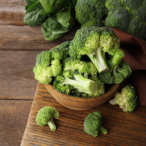 A bowl of Broccoli in a wooden bowl over a wooden tabletop with other green vegetables in the background.